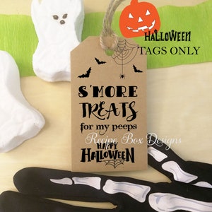 May include: A brown paper tag with the text "S'MORE TREATS for my peeps HAPPY HALLOWEEN" and a spiderweb design. The tag is adorned with a jute string and a pumpkin illustration. White ghost-shaped marshmallows and a black skeleton hand are also visible.