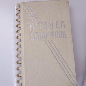 May include: A beige, spiral-bound book with the title "Kitchen Scrapbook" printed in silver lettering. The cover has a textured surface and a diagonal design of silver lines.