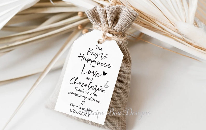 Puede incluir: Bolsa de regalo blanca con una etiqueta que dice "La clave de la felicidad es el amor y los chocolates." Gracias por celebrar con nosotros. Dennis & Allie 02/17/202X