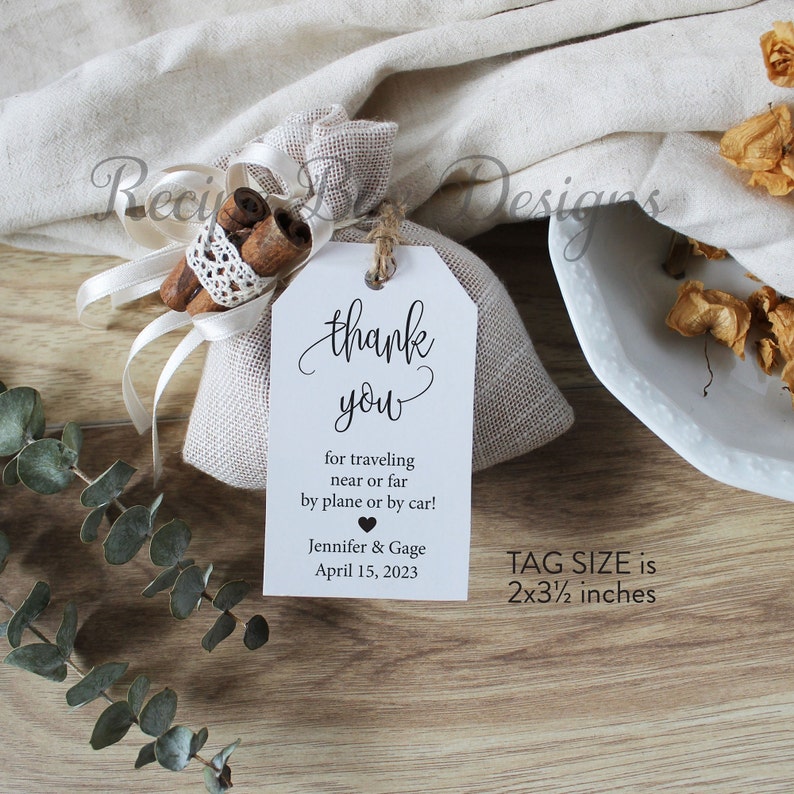 May include: A white thank you tag with black text that reads "thank you for traveling near or far by plane or by car! Jennifer & Gage April 15, 2023". The tag is attached to a small burlap bag with cinnamon sticks inside. The tag size is 2x3 1/2 inches.