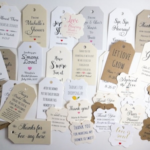 May include: A collection of assorted gift tags in various shapes and sizes. The tags feature handwritten calligraphy and printed text, with phrases like "Thank You," "Love is Sweet," and names with dates. The tags are in shades of white, cream, and brown.