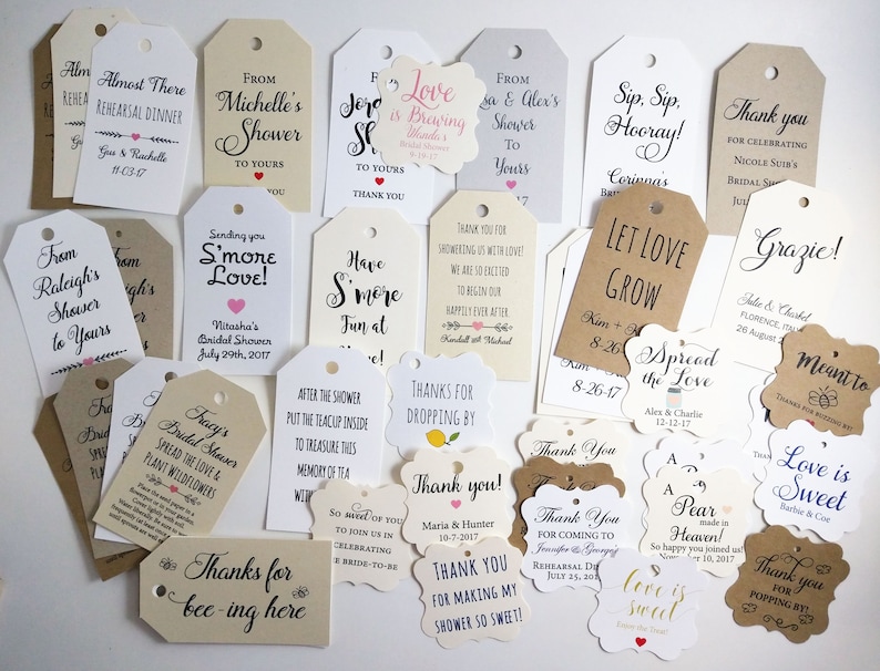May include: A collection of assorted gift tags in various shapes and sizes. The tags feature handwritten calligraphy and printed text, with phrases like "Thank You," "Love is Sweet," and names with dates. The tags are in shades of white, cream, and brown.