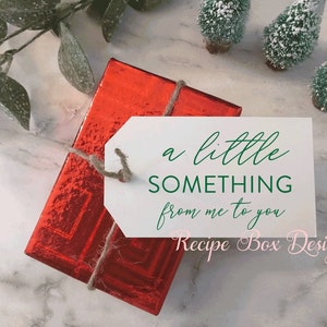 K&ouml;nnte beinhalten: Eine rechteckige Geschenkbox, eingewickelt in gl&auml;nzendes rotes Metallic-Papier, mit einem wei&szlig;en Geschenkanh&auml;nger aus Papier, der mit einer Schnur befestigt ist. Der Anh&auml;nger tr&auml;gt gr&uuml;nen Text a little SOMETHING from me to you und rosa Text Recipe Box Designs.