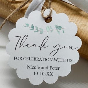 May include: A white scalloped-edge gift tag with the words "Thank you" in elegant script. The tag includes the text "FOR CELEBRATION WITH US!" and the names "Nicole and Peter" with a date. The tag is tied with twine and a white ribbon.