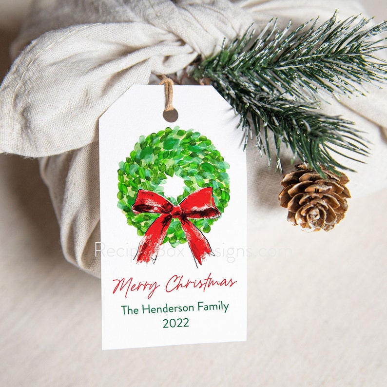 May include: A white gift tag with a watercolor wreath and red bow, with the text "Merry Christmas" and "The Henderson Family 2022". The tag is tied to a wrapped gift with a sprig of pine and a pinecone.