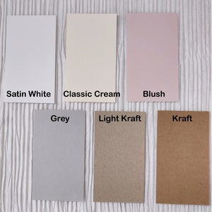 May include: A collection of rectangular paper samples in various colors, including Satin White, Classic Cream, Blush, Grey, Light Kraft, and Kraft. Each sample has a label with its color name. The papers are arranged on a textured white surface.