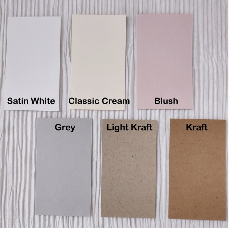 May include: A collection of rectangular paper samples in various colors, including Satin White, Classic Cream, Blush, Grey, Light Kraft, and Kraft. Each sample has a label with its color name. The papers are arranged on a textured white surface.
