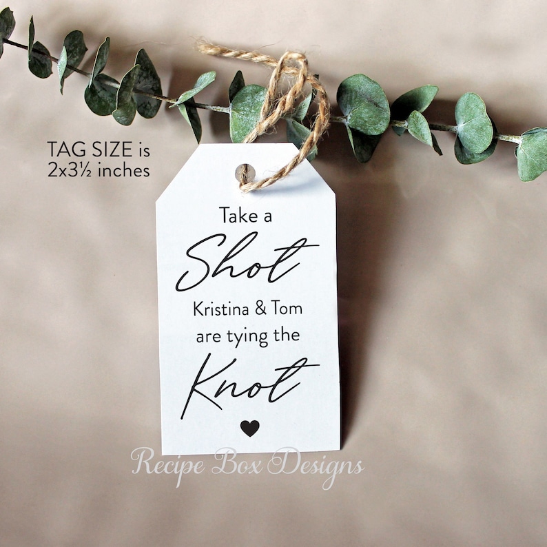 May include: White wedding favor tag with black text that reads "Take a Shot Kristina & Tom are tying the Knot" with a small black heart at the bottom. The tag is 2 by 3 1/2 inches.