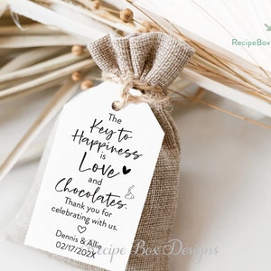 Puede incluir: Una etiqueta de regalo blanca con texto negro que dice "The Key to Happiness is Love and Chocolates." La etiqueta est&aacute; atada a una peque&ntilde;a bolsa de arpillera. El texto tambi&eacute;n incluye "Thank you for celebrating with us. Dennis & Allie 02/17/202X"