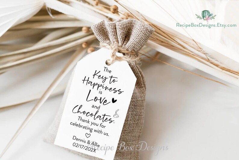 Puede incluir: Una etiqueta de regalo blanca con texto negro que dice "The Key to Happiness is Love and Chocolates." La etiqueta est&aacute; atada a una peque&ntilde;a bolsa de arpillera. El texto tambi&eacute;n incluye "Thank you for celebrating with us. Dennis & Allie 02/17/202X"