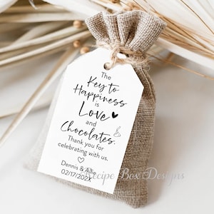 Puede incluir: Una etiqueta de regalo blanca con el texto "The Key to Happiness is Love and Chocolates."  Gracias por celebrar con nosotros. Dennis & Allie 02/17/202X. La etiqueta est&aacute; atada a una peque&ntilde;a bolsa de arpillera.