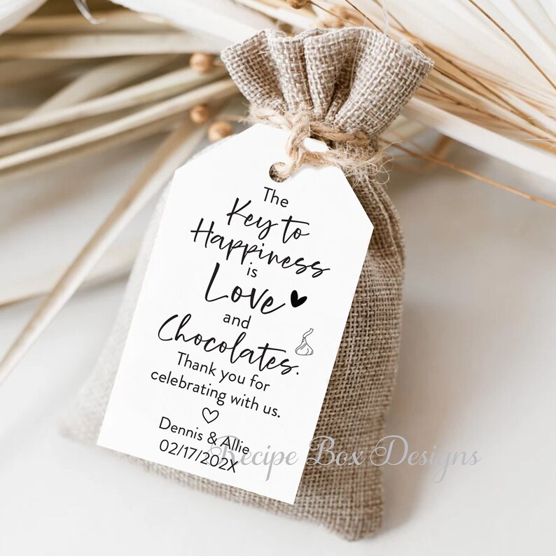 Puede incluir: Una etiqueta de regalo blanca con el texto "The Key to Happiness is Love and Chocolates."  Gracias por celebrar con nosotros. Dennis & Allie 02/17/202X. La etiqueta est&aacute; atada a una peque&ntilde;a bolsa de arpillera.