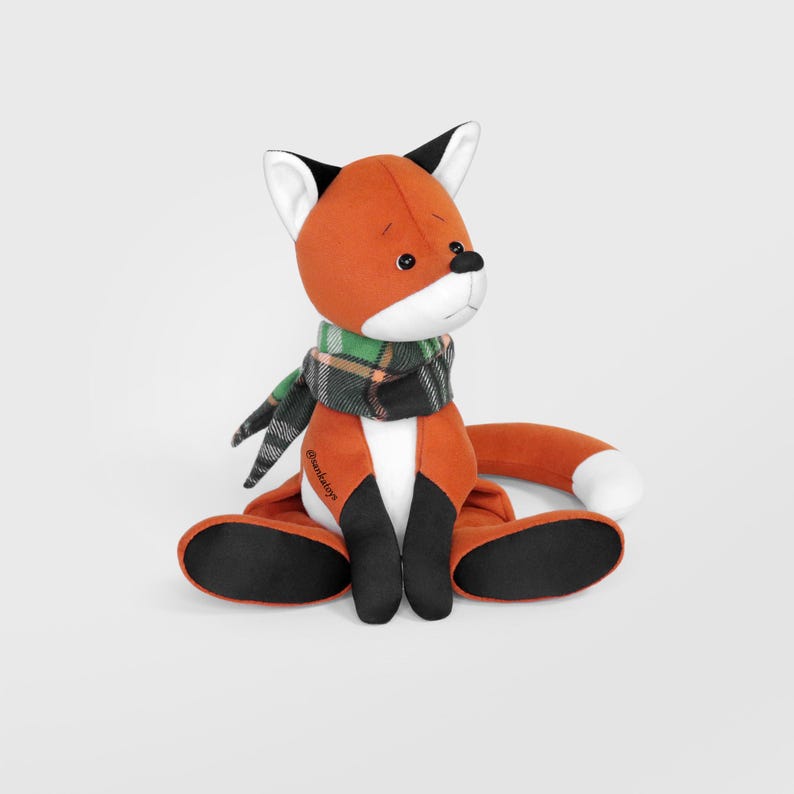 Op de afbeelding: Een pluchen vos met oranje vacht, zwarte poten en een witgepunte staart. Hij draagt een groen-zwarte geruite sjaal. De vos zit met uitgestrekte benen. Het speelgoed heeft een zwarte neus en kleine zwarte ogen.