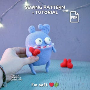 Puede incluir: Un peluche de monstruo de fieltro azul claro con un corazón rojo, con ojos blancos y una sonrisa con dientes. La imagen incluye el texto "SEWING PATTERN + TUTORIAL" y "I'm soft". Un icono PDF está en la esquina superior derecha.