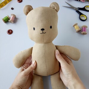 May include: A handmade tan teddy bear doll with a stitched face, small black eyes, and a stitched smile. The doll is being held by two hands. Sewing supplies, including buttons, thread spools, and a measuring tape, are in the background.