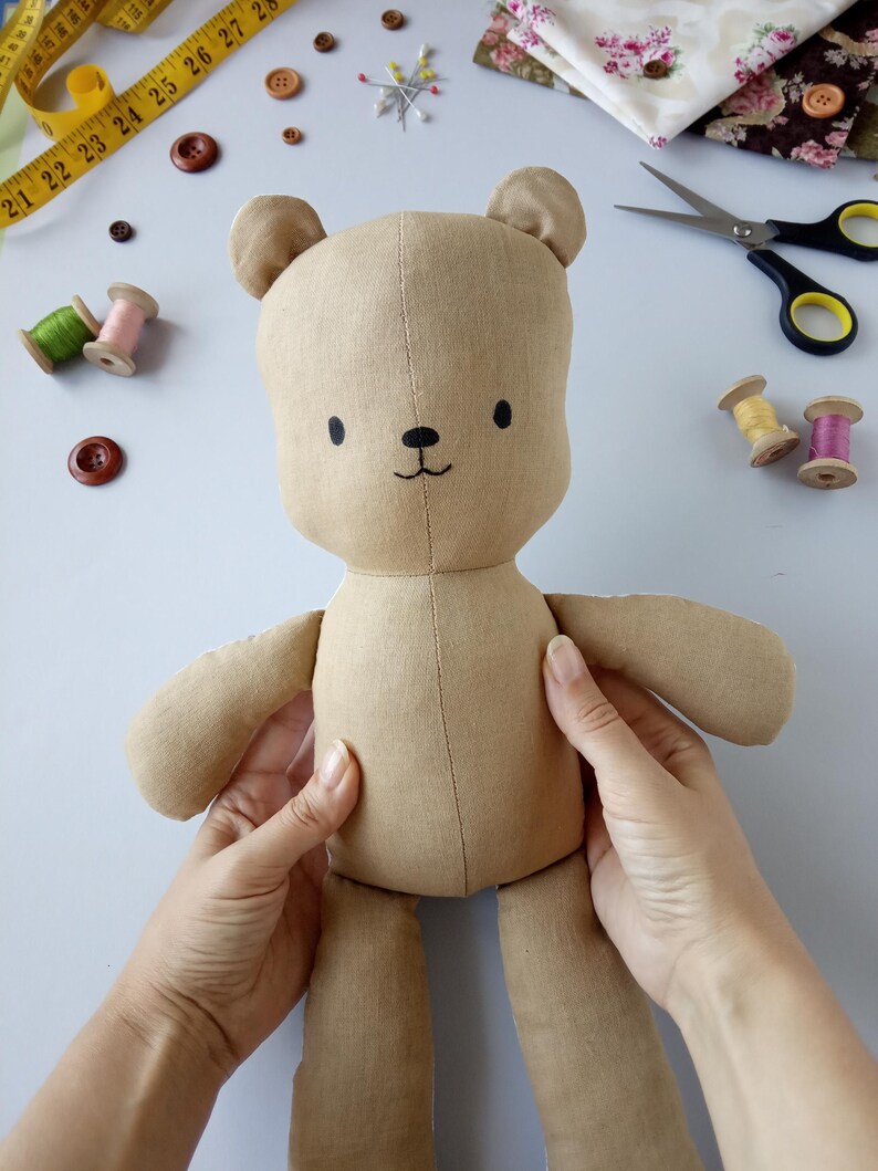 May include: A handmade tan teddy bear doll with a stitched face, small black eyes, and a stitched smile. The doll is being held by two hands. Sewing supplies, including buttons, thread spools, and a measuring tape, are in the background.
