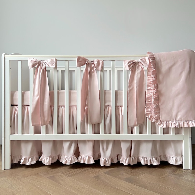A white crib with pink ruffled bedding and bows on the rail.