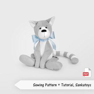 Peut inclure: Un chat en peluche gris et blanc, orné d'un nœud bleu. Le chat a une queue rayée et un nez noir. L'image comprend le texte "Sewing Pattern + Tutorial, Sankatoys".