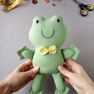 Può includere: Un peluche a forma di rana verde fatto a mano con un papillon giallo. La rana ha occhi neri a bottone e un sorriso cucito. Il peluche è tenuto in mano. Forniture per cucito sullo sfondo.
