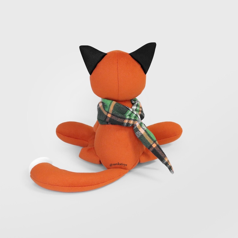 Op de afbeelding: Een oranje pluche vos speelgoed met zwarte oren en een wit puntje aan de staart. De vos draagt een groen, zwart en oranje geruite sjaal. Het speelgoed zit, met de armen en benen uitgestrekt. De tekst "@sankatoys" is zichtbaar op het lichaam van de vos.