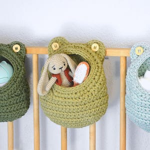 Può includere: Tre cesti portaoggetti fatti a mano a forma di rana all'uncinetto in tonalità di verde e blu sono appesi a una culla di legno. Un cesto contiene un coniglietto di peluche con un gilet rosso. I cesti sono adornati con bottoni di legno.