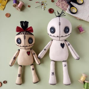 May include: Two handmade rag dolls with button eyes and stitched features. One doll has a red bow and a heart-shaped patch, the other has a black heart. The dolls are beige and white, with black stitching.