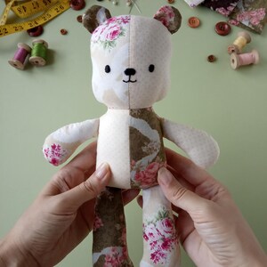 May include: A handmade teddy bear with a floral pattern on one side and solid cream fabric on the other. The bear has embroidered facial features and is being held by a person. Sewing notions, including a measuring tape, spools of thread, and buttons, are scattered around the bear.