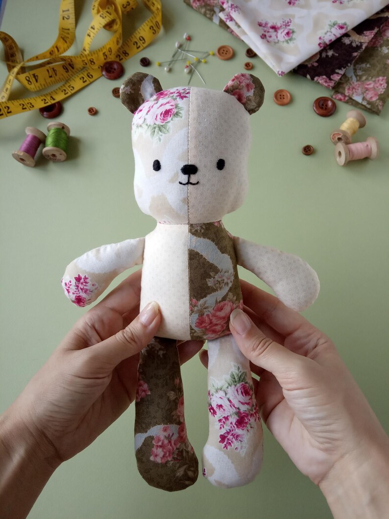 May include: A handmade teddy bear with a floral pattern on one side and solid cream fabric on the other. The bear has embroidered facial features and is being held by a person. Sewing notions, including a measuring tape, spools of thread, and buttons, are scattered around the bear.