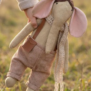 Puede incluir: Un peluche de elefante con un gorro de aviador marrón, una bufanda de ganchillo beige y pantalones de espiga marrones con tirantes. El elefante tiene orejas grandes forradas de rosa, una trompa larga y un pequeño bolsillo en el pantalón.