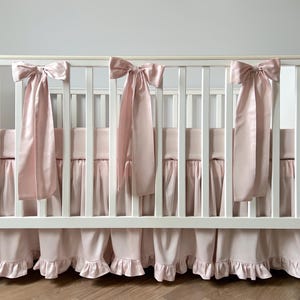 A white crib with pink ruffled bedding and bows on the rail.
