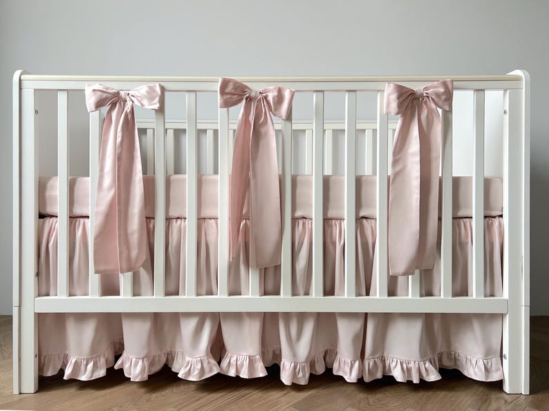 A white crib with pink ruffled bedding and bows on the rail.