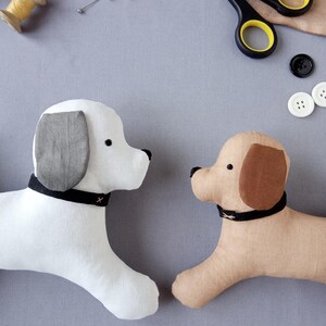 May include: Two plush dog toys, one white and one tan, with black collars and black noses. The white dog has gray ears, and the tan dog has brown ears. Sewing supplies, including scissors and buttons, are in the background.