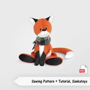 Op de afbeelding: Een oranje pluche vos met een witte borst en zwarte accenten op de oren en voeten. De vos draagt een groen, zwart en wit geruite sjaal. De tekst "Sewing Pattern + Tutorial, Sankatoys" staat onder de vos.