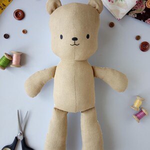 May include: A handmade tan teddy bear with embroidered facial features. The bear is surrounded by sewing supplies, including buttons, spools of thread, fabric scraps, scissors, and a measuring tape. The bear is made of soft fabric and has a simple, charming design.