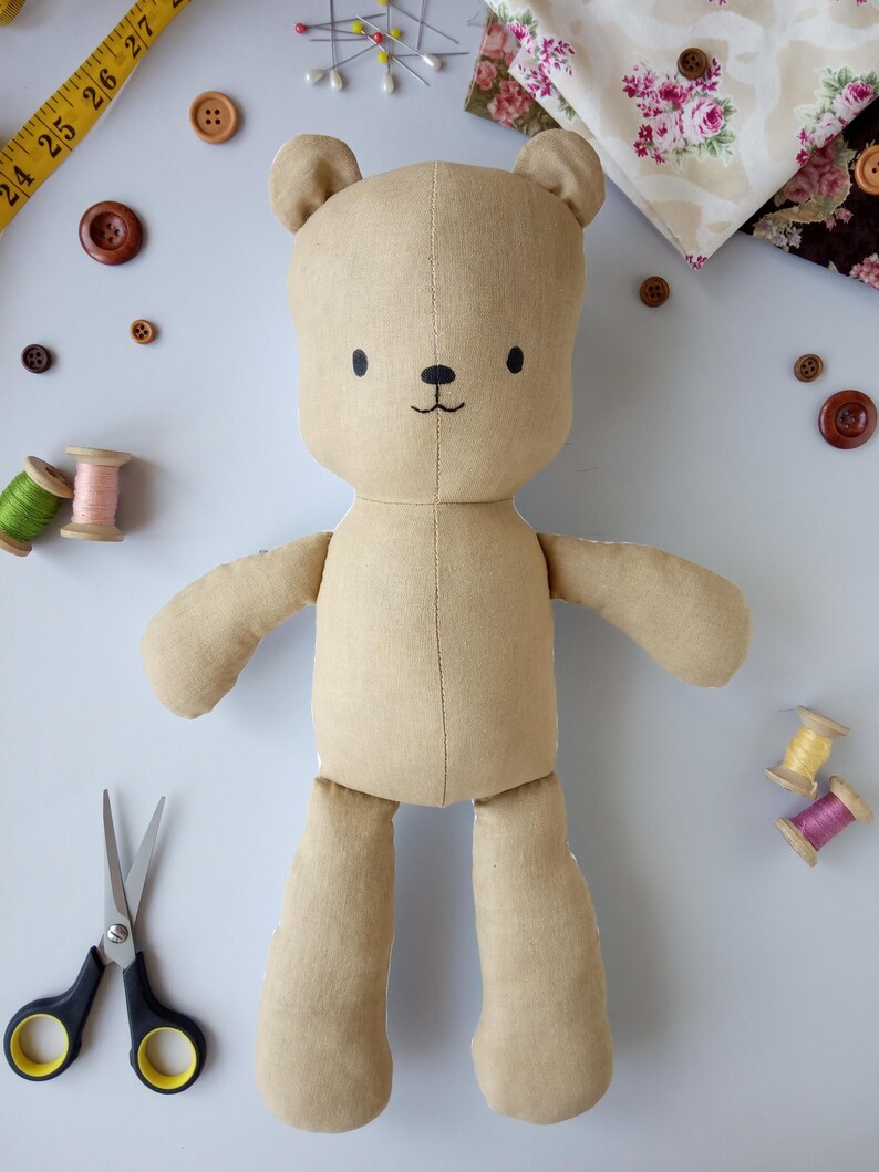 May include: A handmade tan teddy bear with embroidered facial features. The bear is surrounded by sewing supplies, including buttons, spools of thread, fabric scraps, scissors, and a measuring tape. The bear is made of soft fabric and has a simple, charming design.