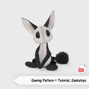 May include: A plush toy with large pointed ears, black and light grey fabric panels, and a fluffy black pom-pom tail. The toy features large black oval eyes and a central seam. Text on the ear reads @sankatoys. Includes a PDF sewing pattern and tutorial.