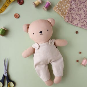 May include: A handmade plush bear doll in a light beige color, wearing a matching jumpsuit. The bear has a black embroidered nose and eyes. Sewing supplies, including spools of thread, buttons, and scissors, are arranged around the doll on a light green surface.