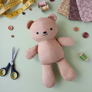 May include: A tan teddy bear with black button eyes and a stitched smile, surrounded by sewing supplies. These include a yellow measuring tape, scissors, wooden buttons, fabric scraps, and spools of thread. The background is a light green.