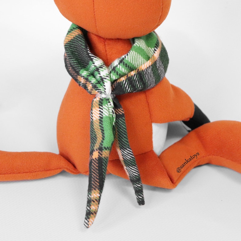 Op de afbeelding: Een oranje pluche vos speelgoed met een groen, zwart en wit geruite sjaal om de nek geknoopt. De vos heeft zwarte accenten op zijn oren en voeten. Het speelgoed zit met uitgestrekte benen.