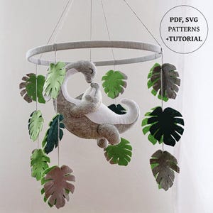May include: A hanging mobile featuring a felt dinosaur parent and baby in gray and white, surrounded by various green and brown felt monstera leaves suspended from a circular frame. The image includes text stating PDF, SVG PATTERNS +TUTORIAL.