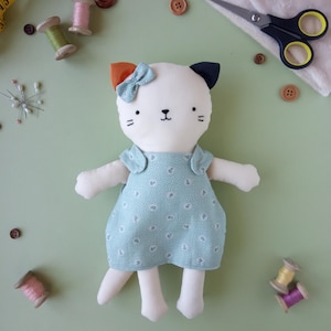 May include: A handmade white cat doll with a blue floral dress and a bow. The doll has orange and navy blue ears, black whiskers, and a stitched face. Sewing supplies, including spools of thread, buttons, and scissors, are scattered around the doll.