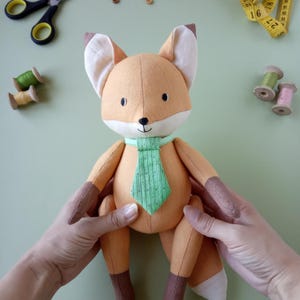 Puede incluir: Muñeco de peluche de zorro hecho a mano, con tela naranja, marrón y crema. El zorro lleva una corbata verde con un patrón de rayas verticales. El muñeco está sostenido por manos, con útiles de costura en el fondo.