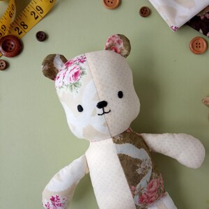 May include: A handmade teddy bear with a floral pattern on its ears, arms, and legs. The bear is made of cream and brown fabric and has a stitched black mouth and eyes. A yellow measuring tape and brown buttons are scattered around the bear.