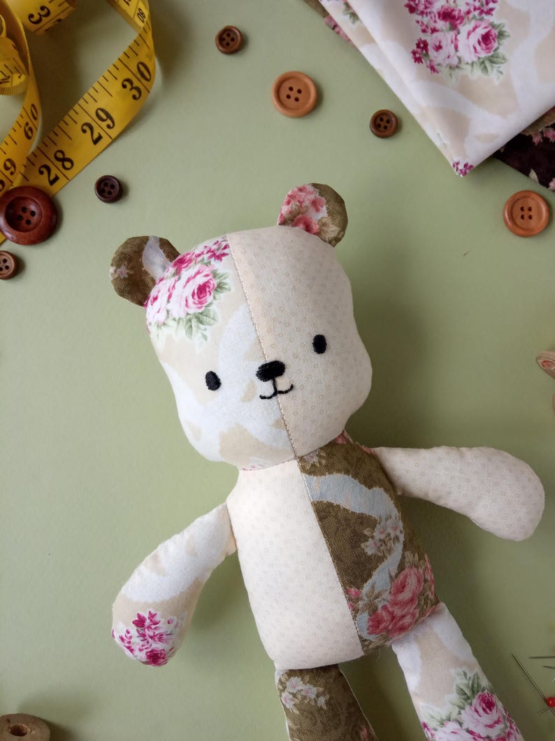 May include: A handmade teddy bear with a floral pattern on its ears, arms, and legs. The bear is made of cream and brown fabric and has a stitched black mouth and eyes. A yellow measuring tape and brown buttons are scattered around the bear.