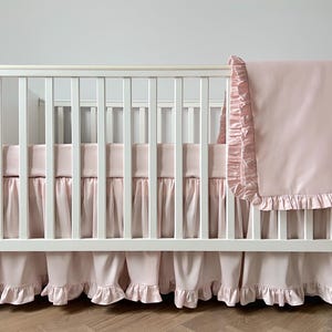 A white wooden crib with a pink blanket and ruffled skirt, placed against a plain white wall.
