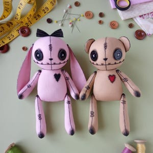 Könnte beinhalten: Zwei handgefertigte Plüschpuppen: ein rosa Hase mit schwarzer Schleife und ein beigefarbener Bär. Beide haben aufgenähte Merkmale, Knopfaugen und herzförmige Aufnäher. Der Hase hat lange Ohren, und der Bär hat kleine, abgerundete Ohren.