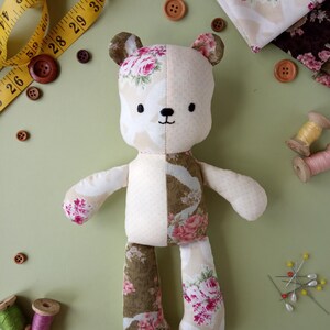 May include: A handmade teddy bear with floral fabric accents. The bear is cream-colored with floral patterned fabric on the ears, arms, and legs. Wooden buttons, measuring tape, fabric, and spools of thread are scattered around the bear on a green surface.