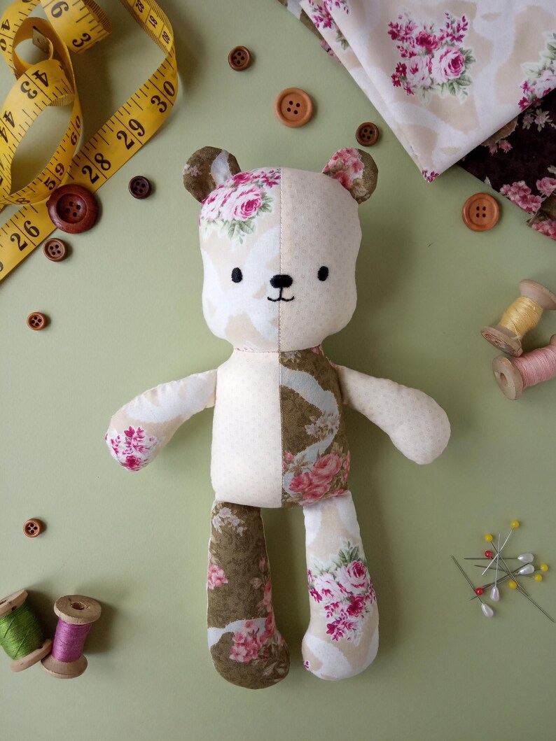 May include: A handmade teddy bear with floral fabric accents. The bear is cream-colored with floral patterned fabric on the ears, arms, and legs. Wooden buttons, measuring tape, fabric, and spools of thread are scattered around the bear on a green surface.