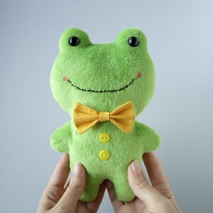 Peut inclure: Une peluche verte en forme de grenouille, ornée d'un nœud papillon jaune et de deux boutons jaunes. La grenouille a des yeux noirs et un sourire cousu. Un jouet doux et câlin pour enfants.