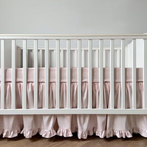 A white wooden crib with a pink mattress cover and ruffled skirt.